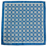 A square blue scarf with a white geometric damask pattern and a solid blue border, featuring the brand name 'A. AZTHOM' printed in white at the bottom left, laid flat on a gray carpet.