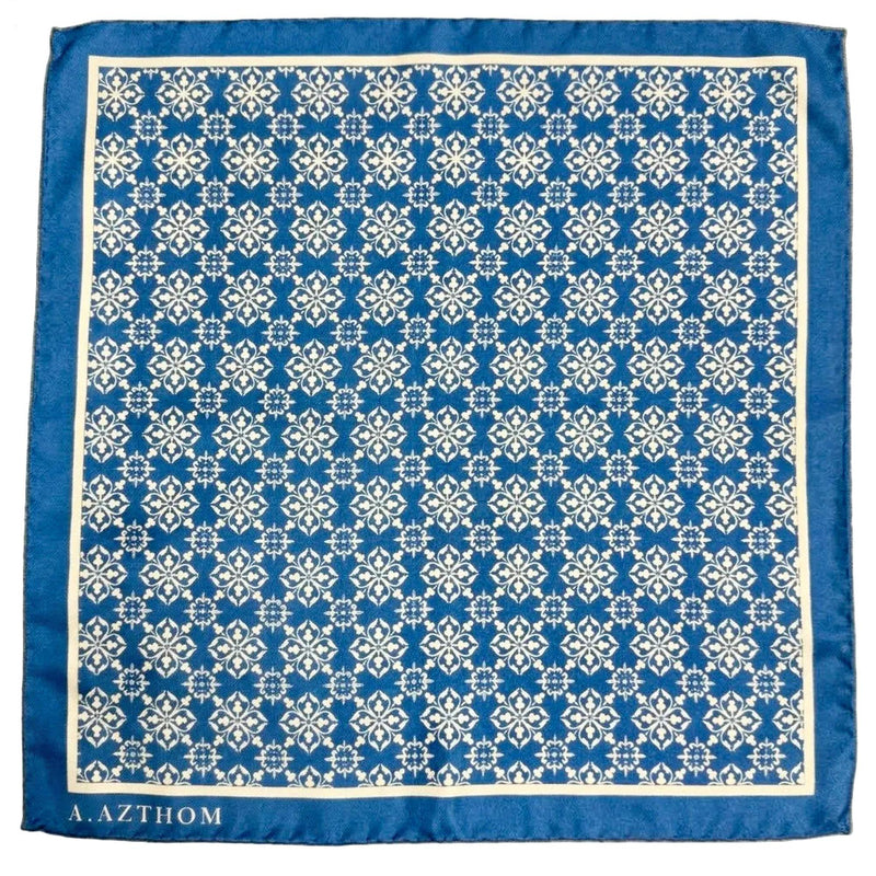 A square blue scarf with a white geometric damask pattern and a solid blue border, featuring the brand name 'A. AZTHOM' printed in white at the bottom left, laid flat on a gray carpet.